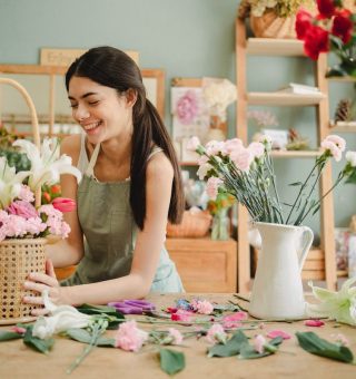 Uma jovem está sorrindo no balcão de uma floricultura enquanto arruma um vaso decorativo