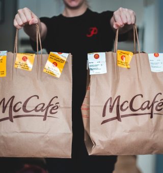 Jovem Aprendiz Mc Donalds segura duas sacolas de comida do McDonald's. As sacolas são vermelhas e amarelas com o logotipo do McDonald's. A pessoa está sorrindo e parece feliz com sua comida.