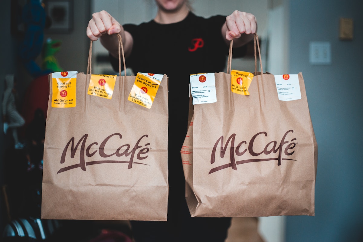 Jovem Aprendiz Mc Donalds segura duas sacolas de comida do McDonald's. As sacolas são vermelhas e amarelas com o logotipo do McDonald's. A pessoa está sorrindo e parece feliz com sua comida.