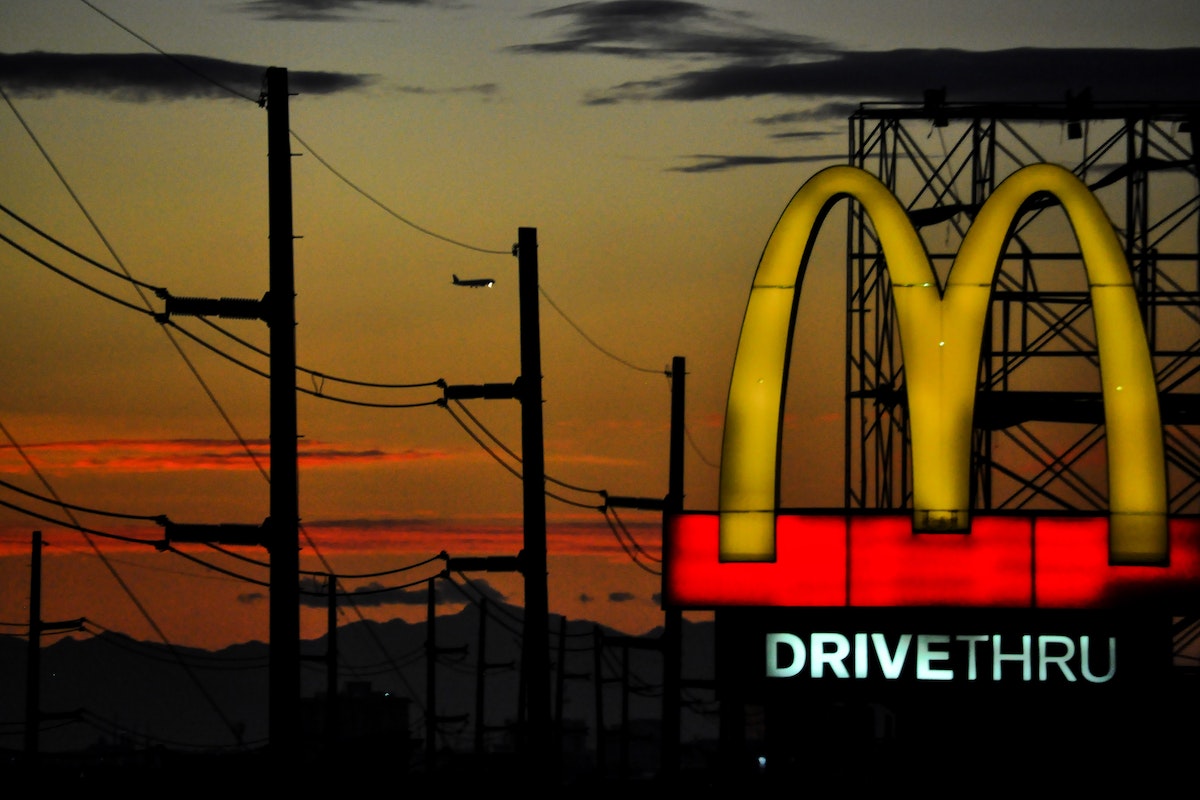 Um letreiro do McDonald's brilha no crepúsculo. O letreiro é vermelho e amarelo e tem o logotipo do McDonald's. O céu atrás do letreiro é laranja e vermelho.