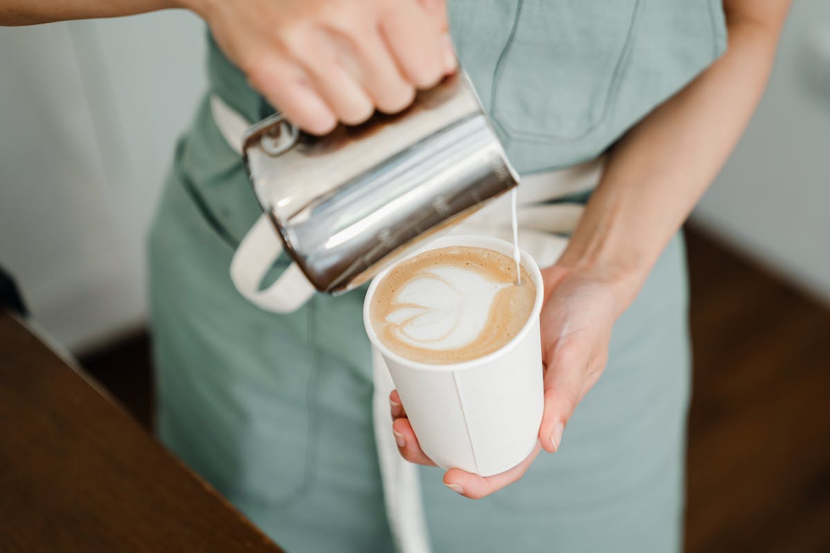 Uma barista serve um café. Ela segura a xícara com a mão e coloca o leite por cima do café na xícara