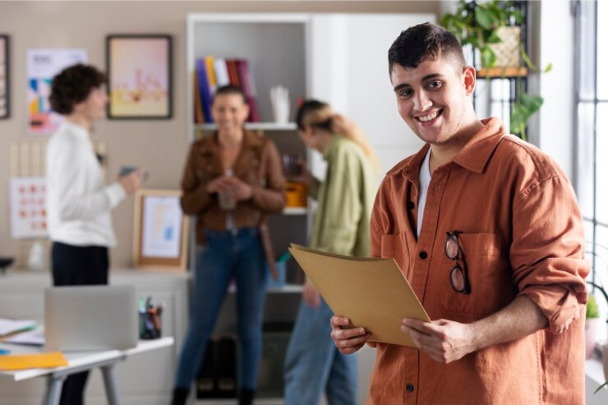 Um homem selecionado pelo Programa Jovem Aprendiz 2024 está sorrindo dentro de um escritório enquanto segura uma pasta