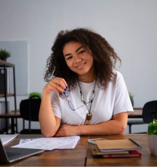 Uma jovem Aprendiz sentada na mesa de um escritório. Ela tem cabelo enrolado e veste blusa branca, e se candidatou pela Rede Cidadã 2024