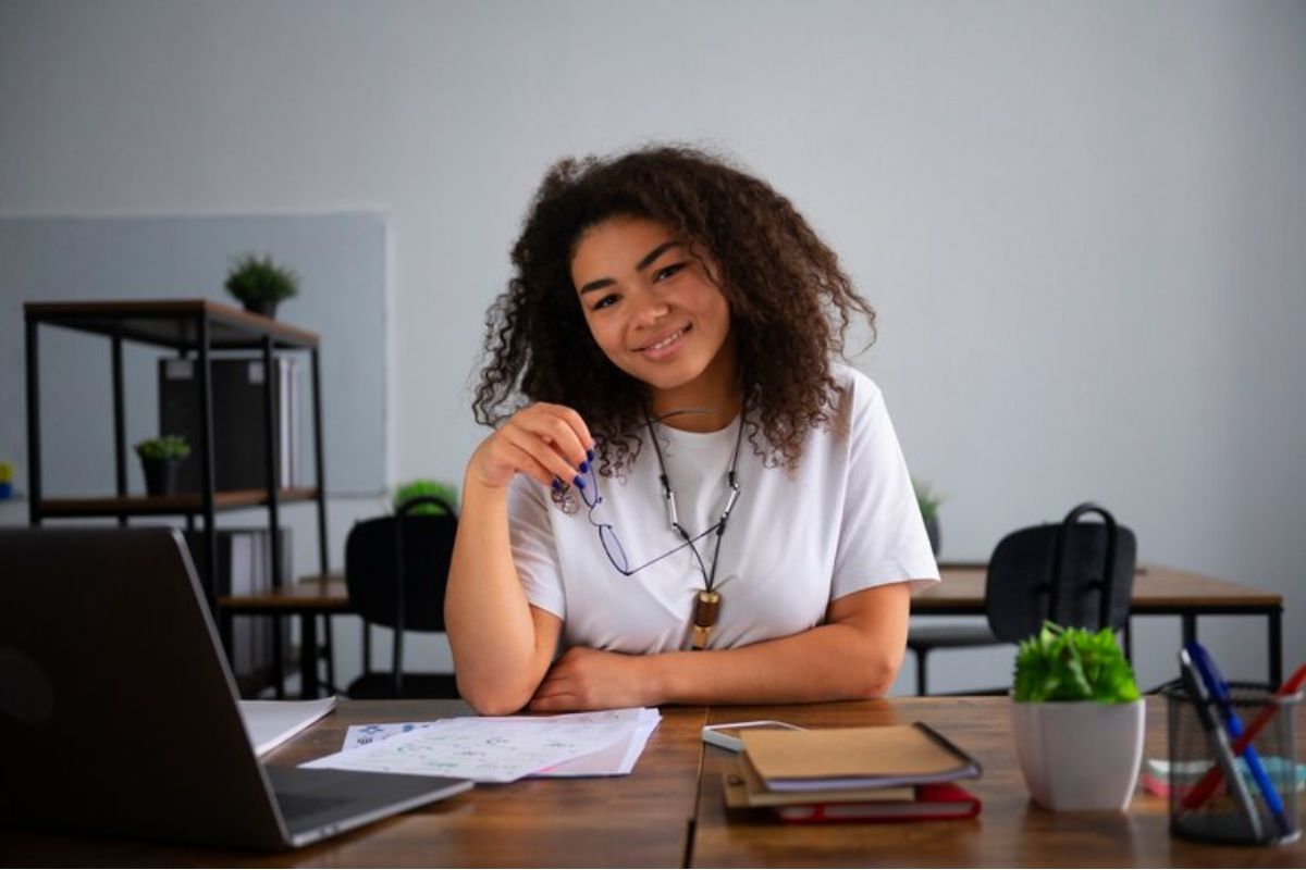 Uma jovem Aprendiz sentada na mesa de um escritório. Ela tem cabelo enrolado e veste blusa branca, e se candidatou pela Rede Cidadã 2024