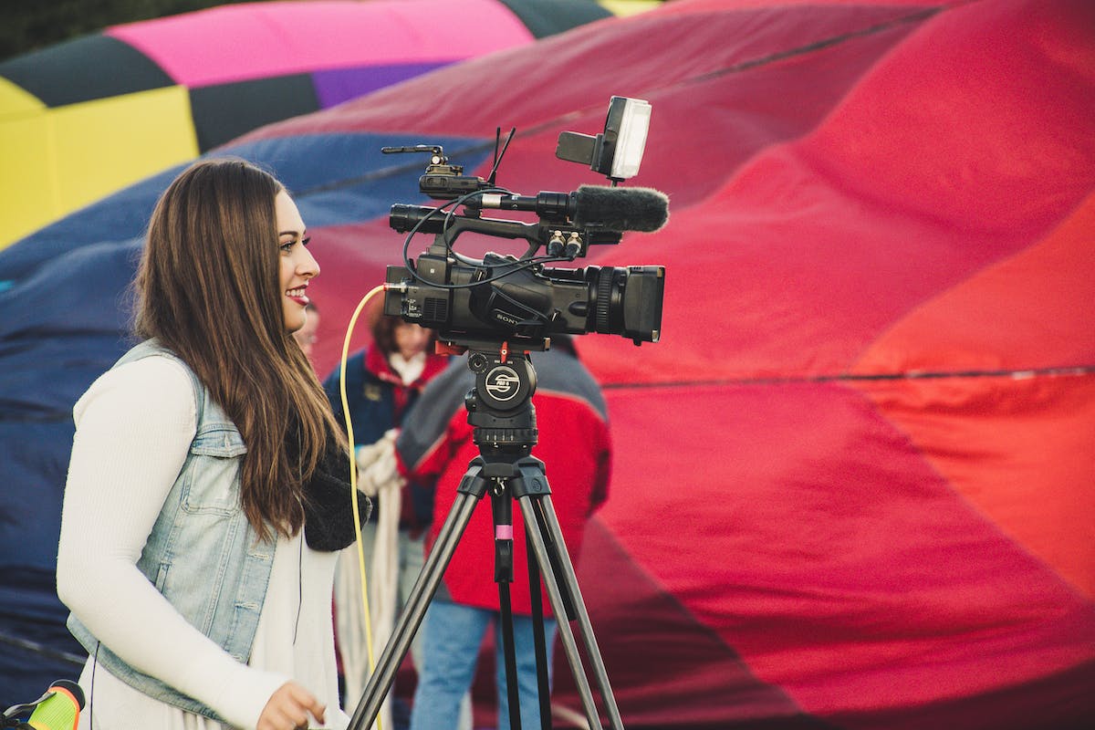 Uma mulher está filmando com uma câmera profissional. Ao fundo tem um tecido vermelho